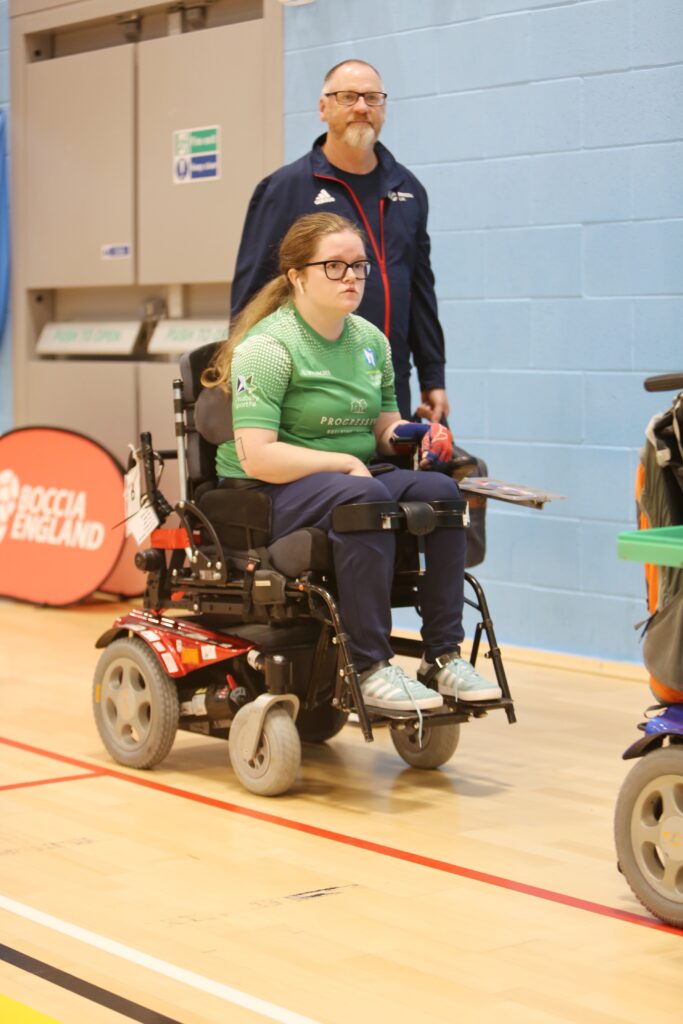 Claire is in her red sports powerchair, not looking at the camera and has earbuds in. Glynn is walking behind her, carrying his handbag