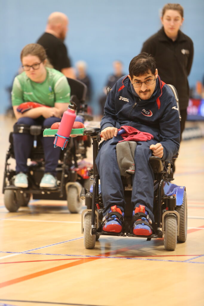 Reshad is in his powerchair lining up to play a shot. He is wearing a navy t shirt and has glasses on. Claire is looking over his shoulder in her powerchair. She is wearing a green t shirt and also wears glasses.