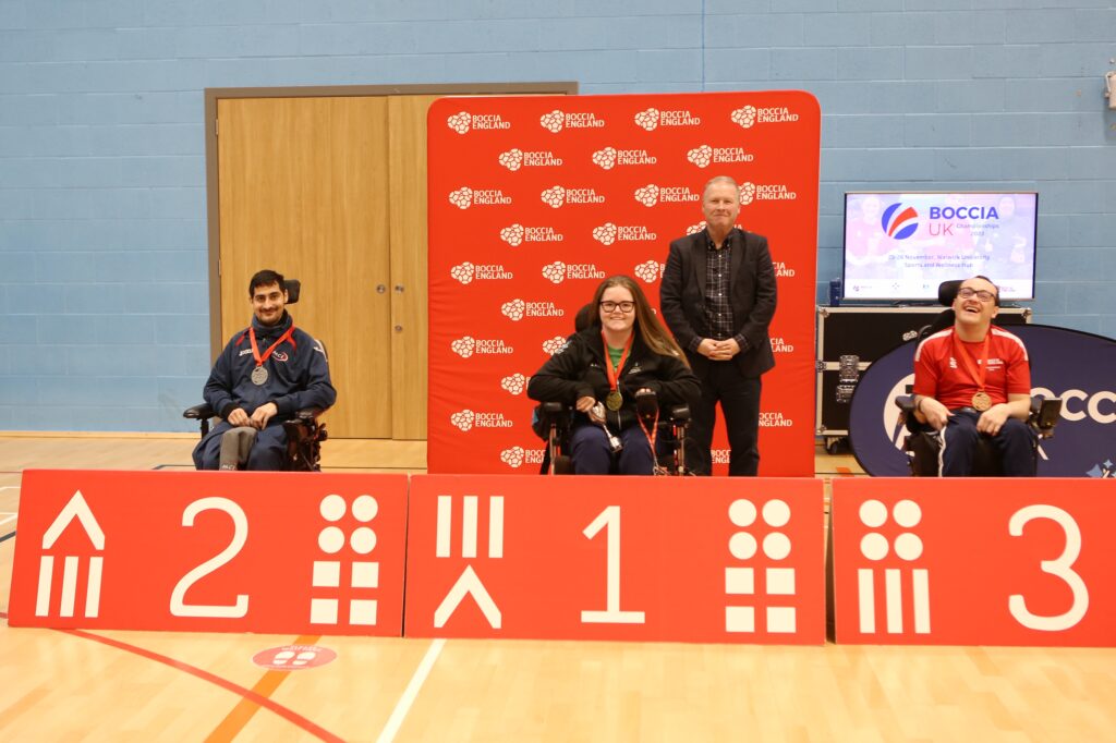 Medal Ceremony for BC2 Classification. Reshad on the left sits behind a board that says 2, Claire is in the middle and sat behind a board that has a 1, James is on the right behind a board with a 3. Jon Morgan, President of World Boccia stands between Claire and James. 