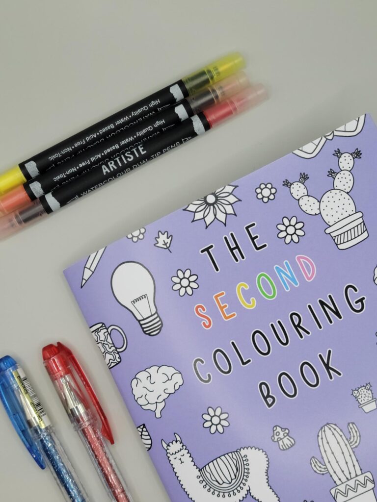 The Second Colouring Book is on the right of the picture. It is purple. 3 felt tip pens are above it, and 2 gel pens are to the left. It is on a grey background.